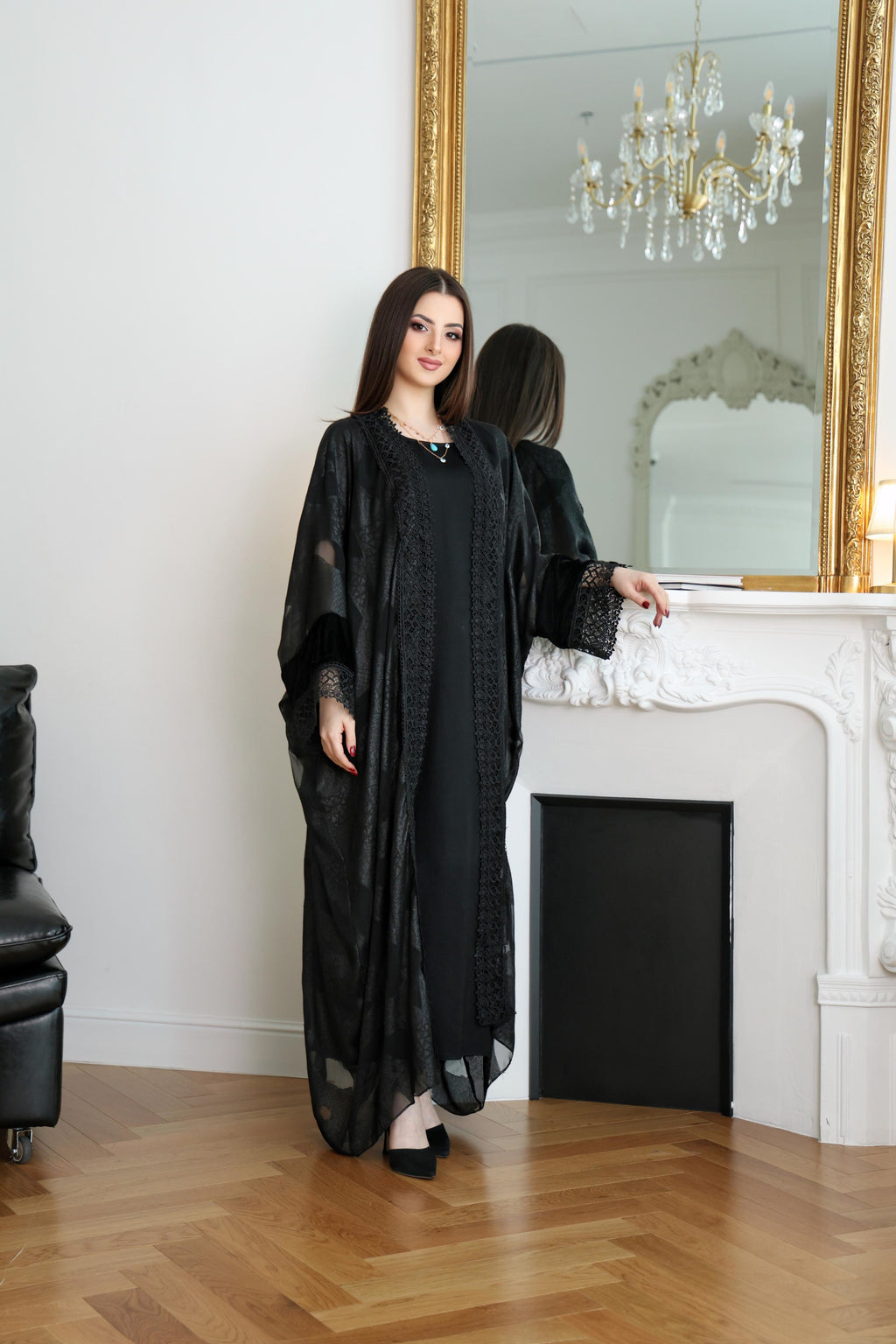 Woman in a black dress standing in a room with a large mirror and chandelier.Black abaya, classic black abaya, luxury abaya, elegant abaya, modest fashion, women’s abaya, Dubai abaya, designer abaya, Eid abaya, Ramadan abaya