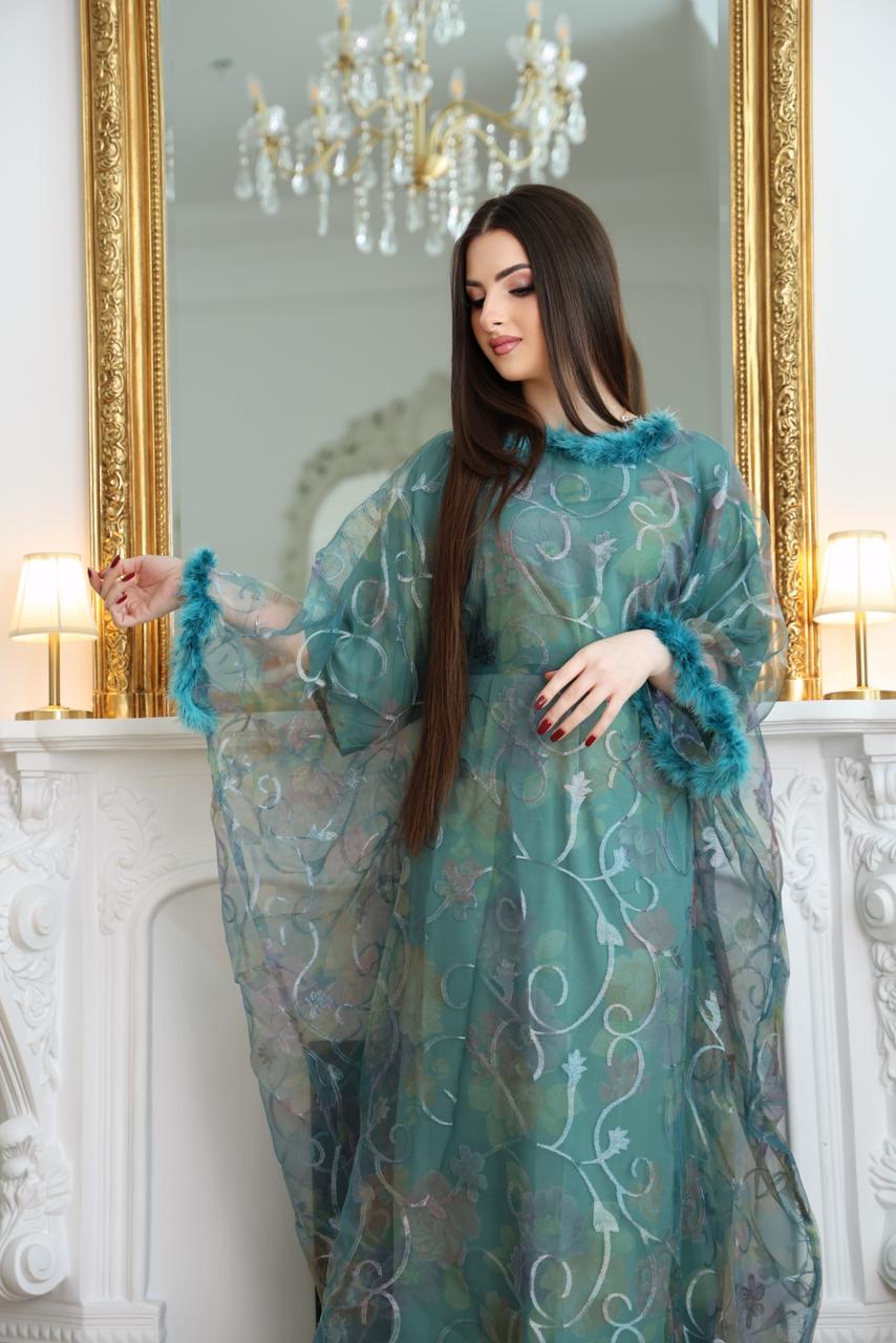Woman wearing a teal floral dress standing in front of a mirror with decorative frames. Light blue abaya, sky blue abaya, elegant abaya, luxury abaya, minimalist abaya, modest fashion, Dubai abaya, Eid abaya, Ramadan abaya