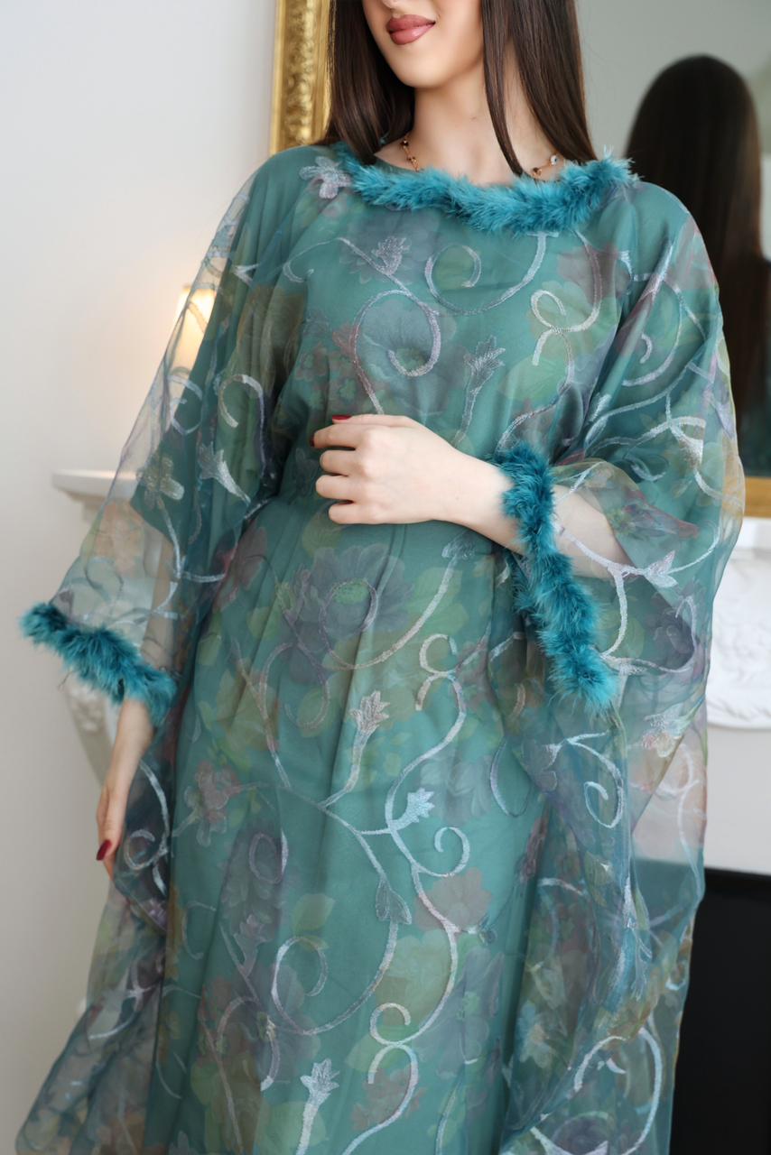 Woman wearing a teal dress with floral patterns and feathered cuffs indoors.