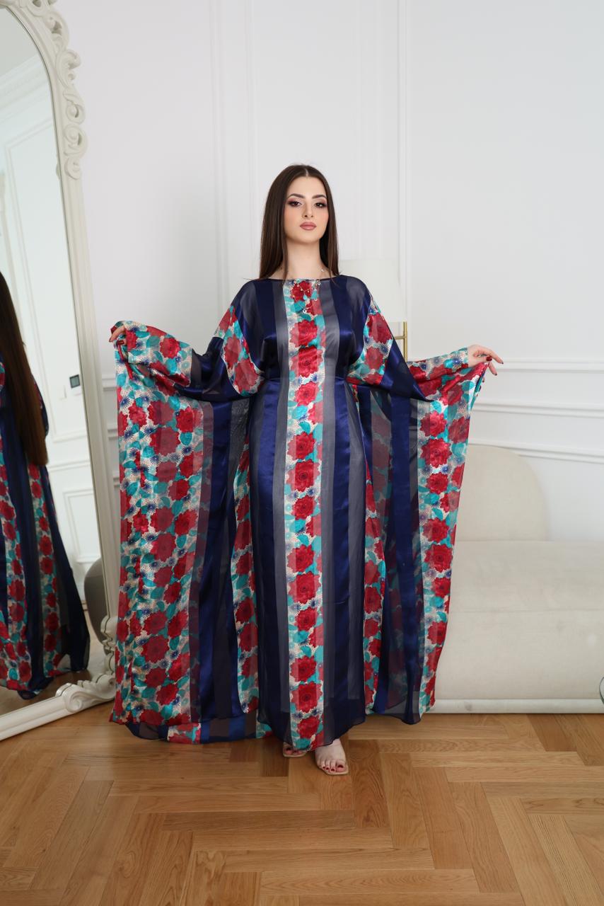 Woman wearing a colorful floral dress standing in a room with a mirror and white wall.Luxury rose abaya with blue and red tones, designed in Dubai. Elegant, lightweight, and perfect for everyday wear and special occasions.
