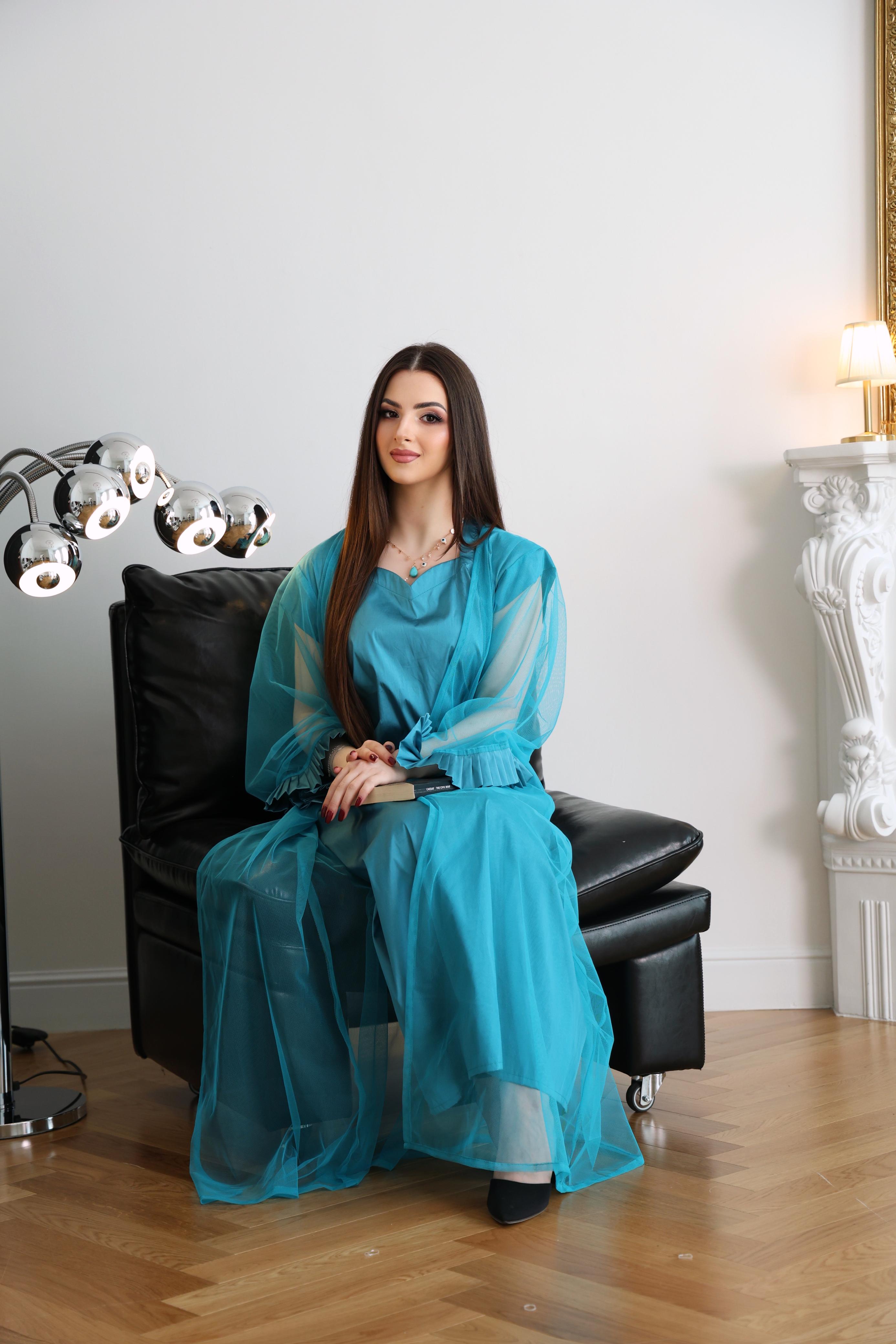 Woman in a blue dress sitting on a black leather chair in a room with white walls and wooden flooring.