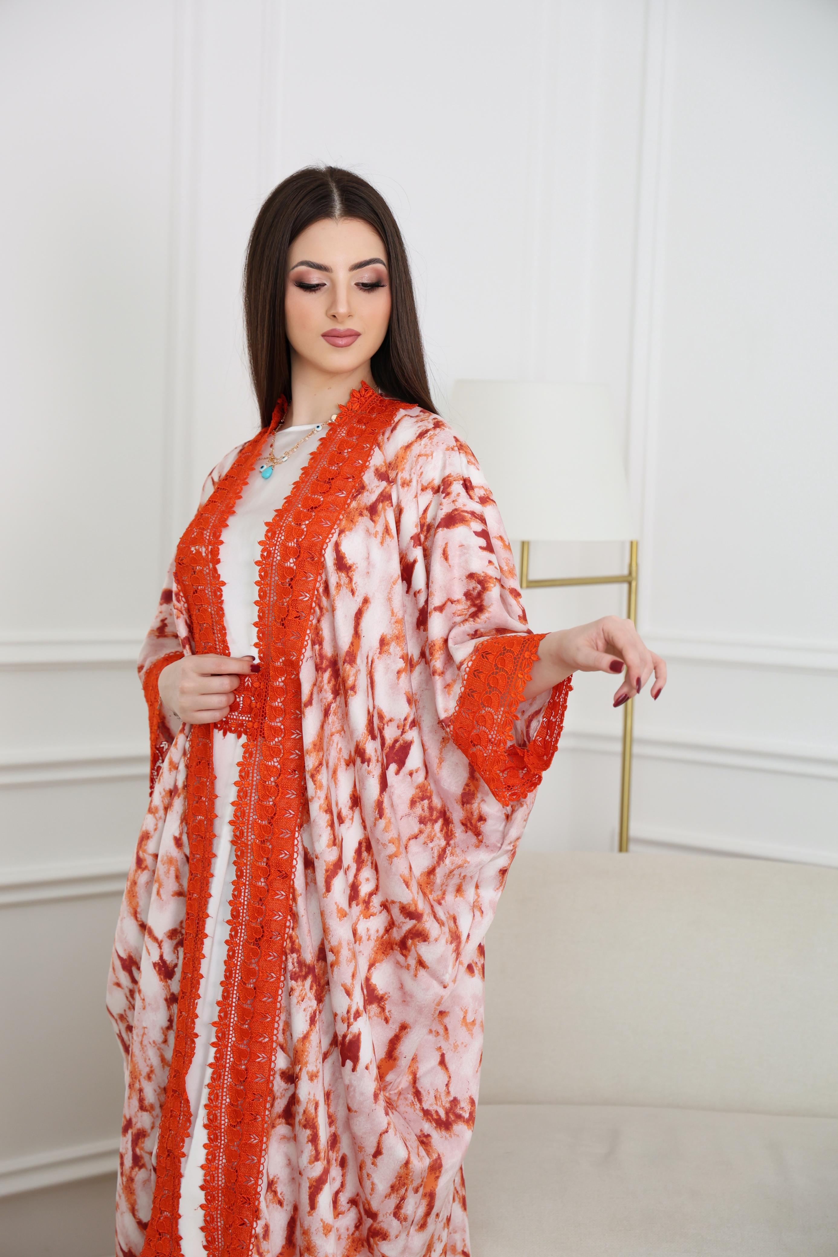 Woman wearing a orange and white patterned Abaya gainst a white wall.