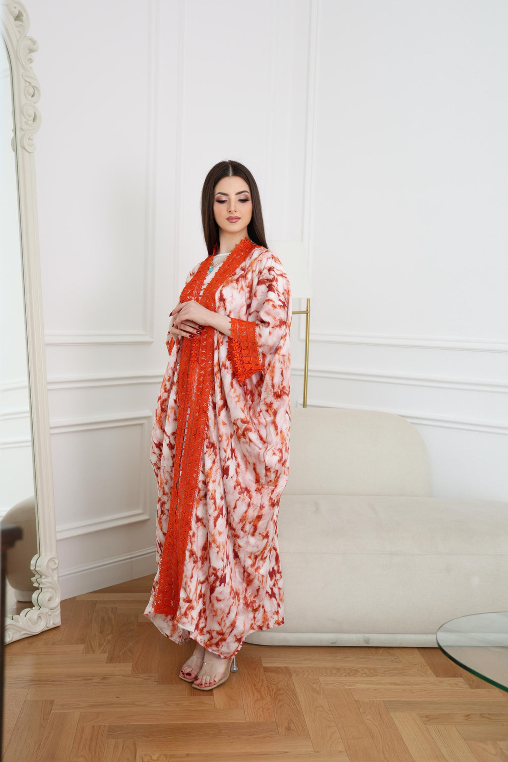 Woman in a floral dress with red accents standing in a room with white walls and wooden floor.