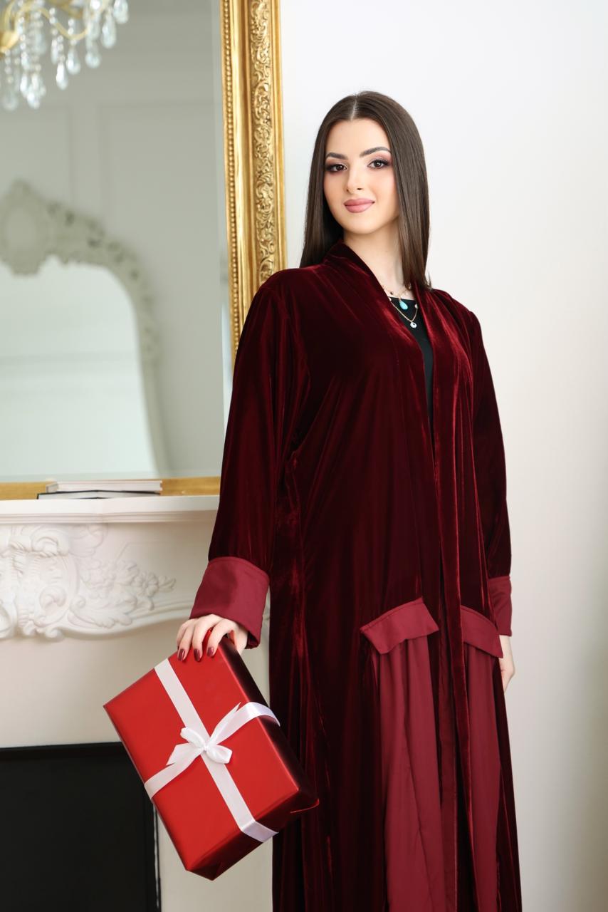 Woman in a burgundy abaya holding a red gift box in a decorated room.