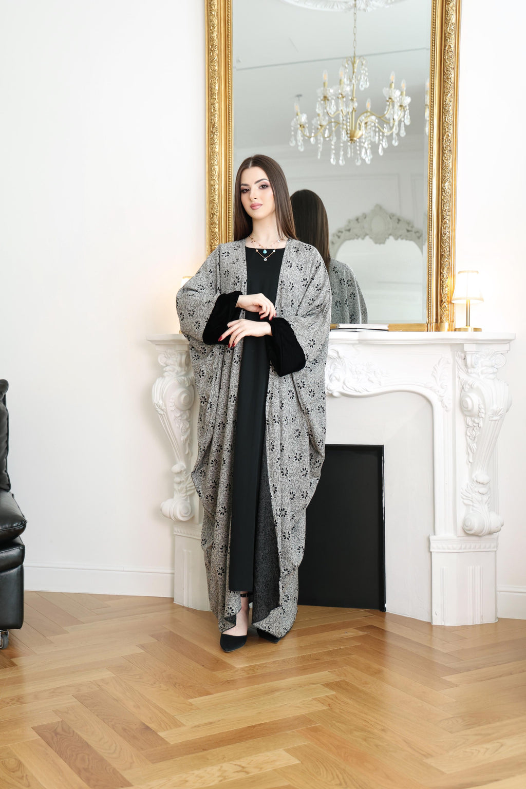 Woman in a gray patterned dress standing in a room with a mirror and chandelier.