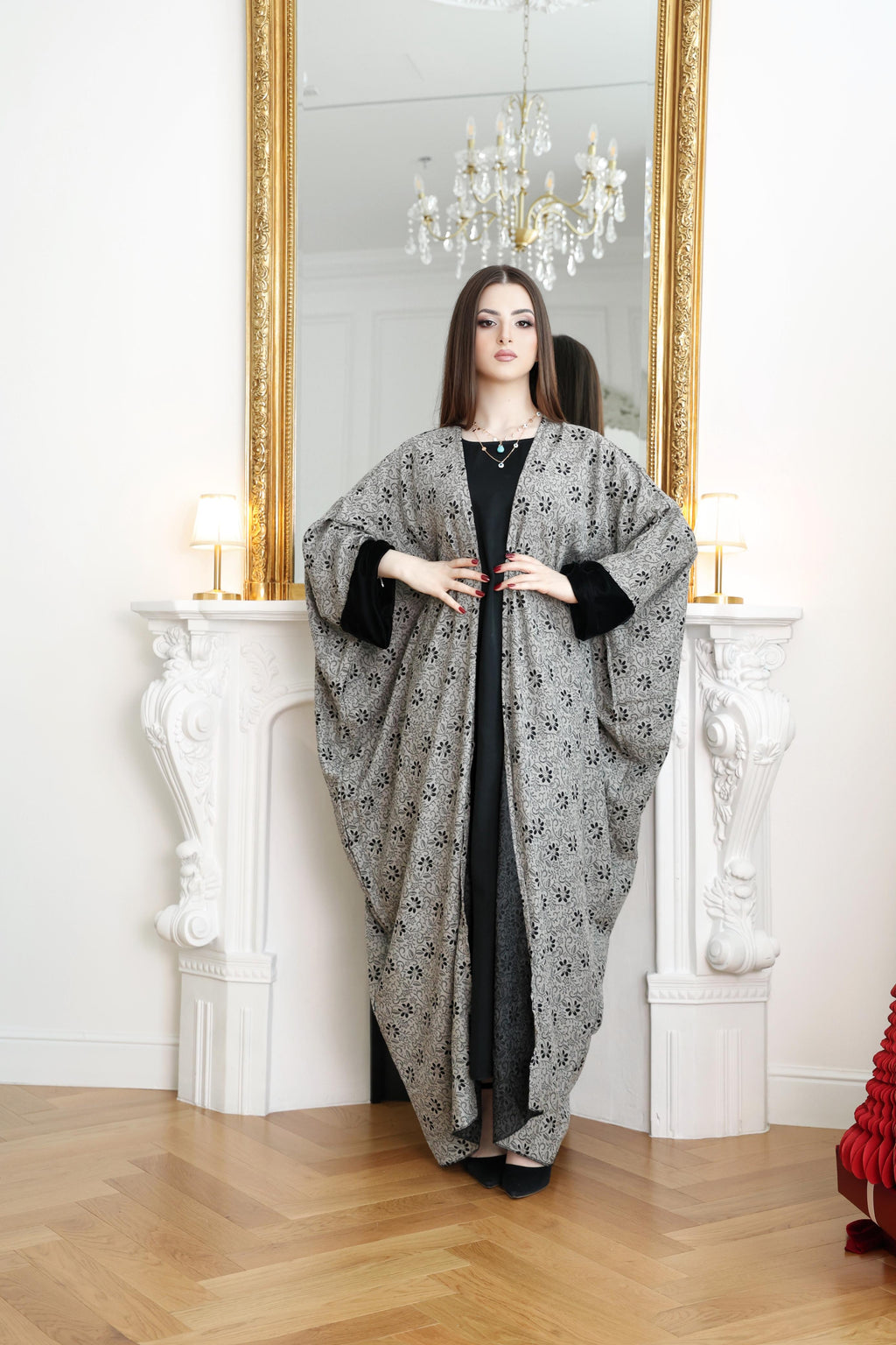 Woman wearing a long, patterned gray shawl over a black outfit in a room with a mirror and chandelier.