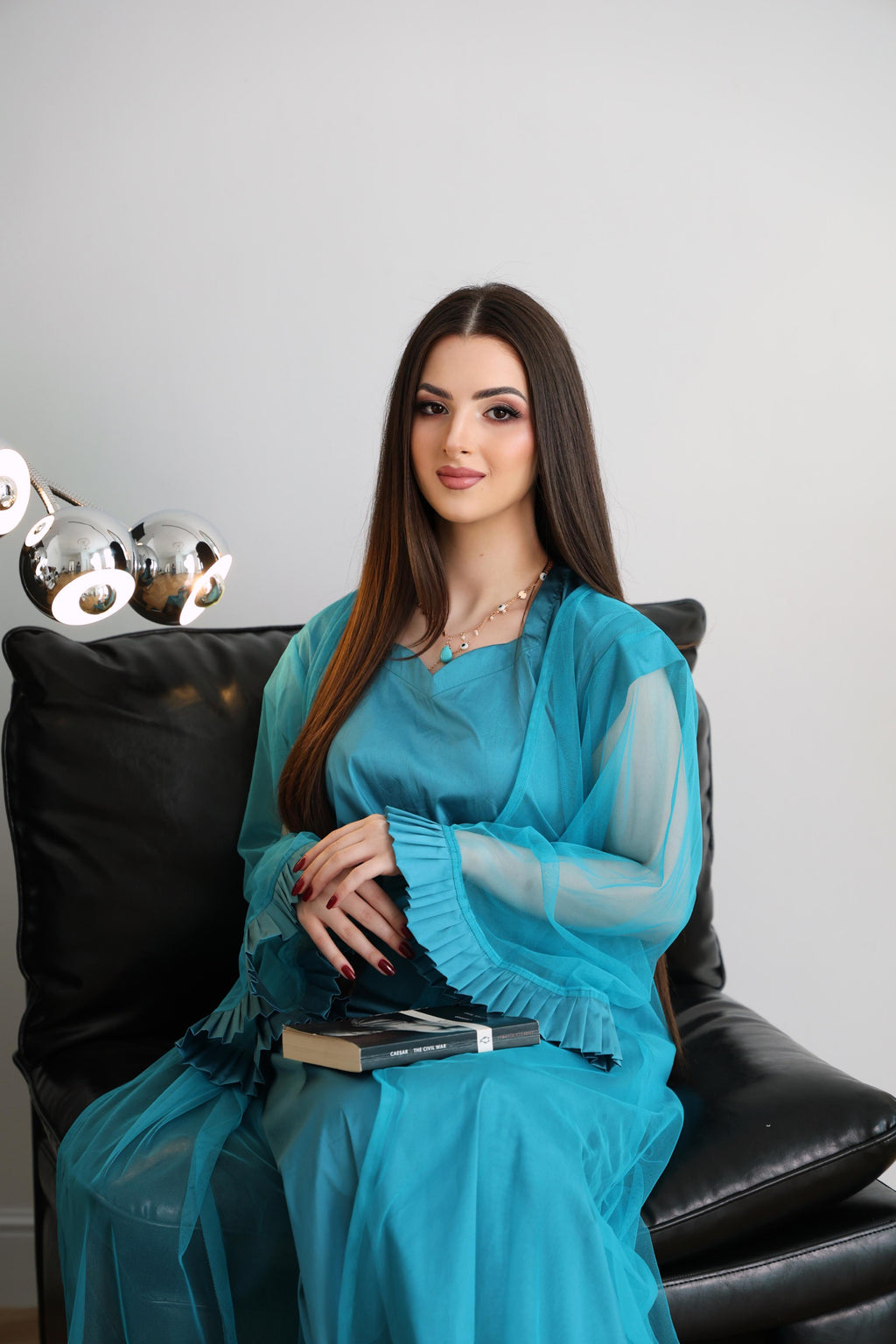 Woman in a blue abaya sitting on a black couch with a white background