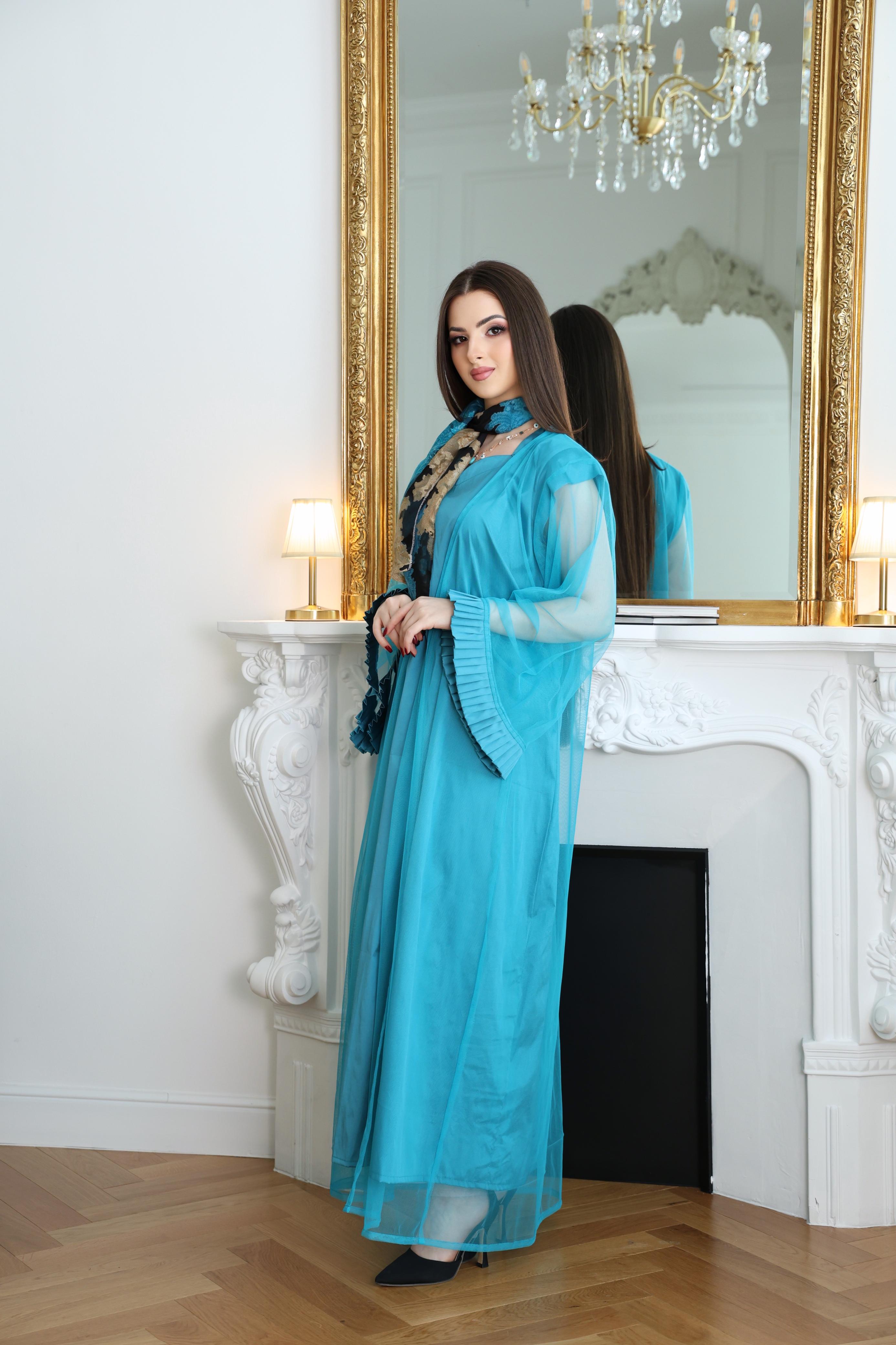 Woman in a turquoise dress standing in front of a mirror in a room with a chandelier.