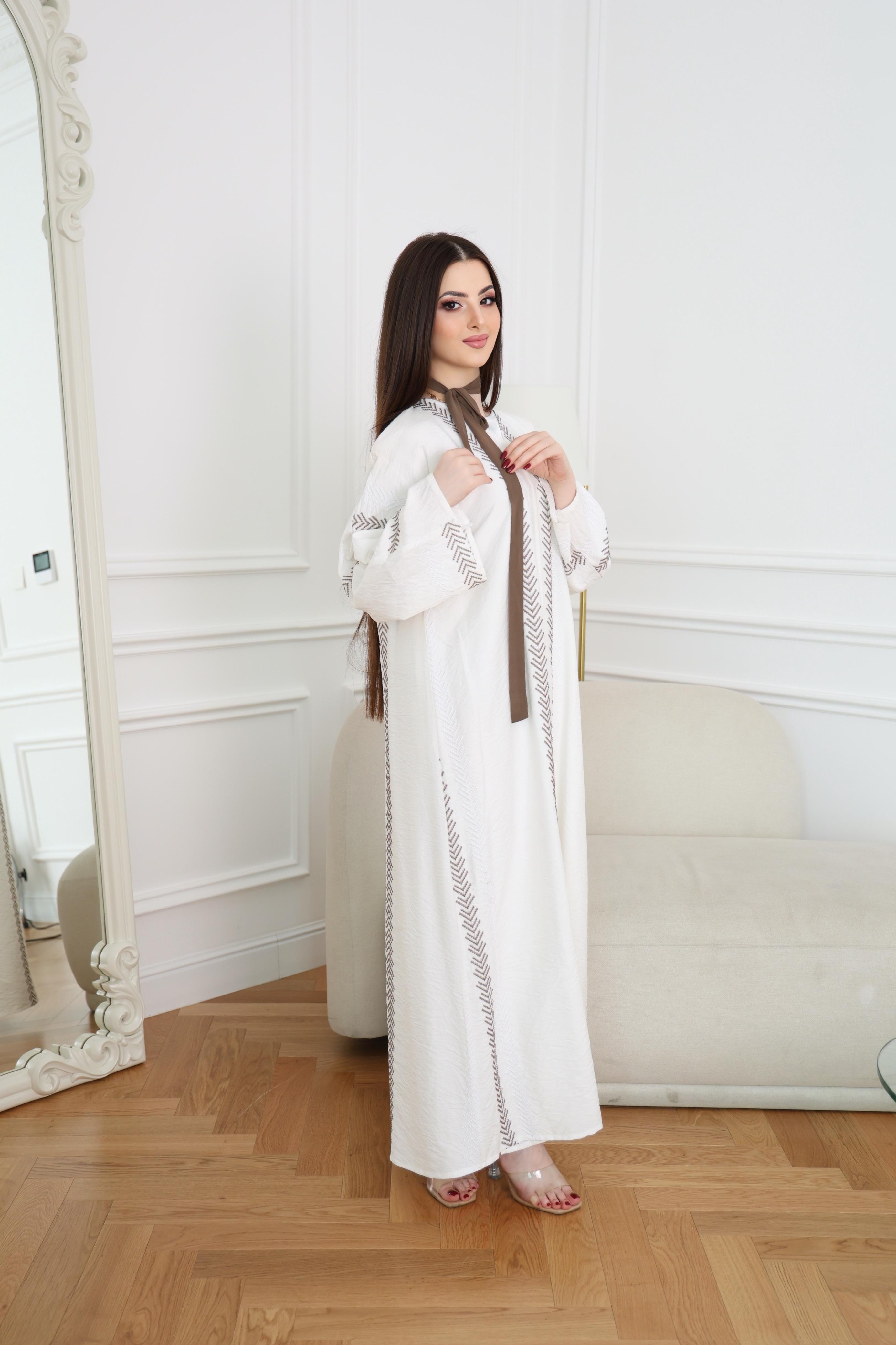 Woman in a white embroidered dress standing in a room with wooden floor and white walls.
